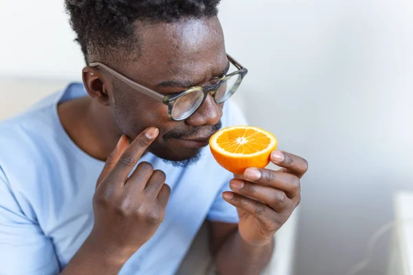 Sick Arican man trying to sense smell of half fresh orange, has symptoms of Covid-19, corona virus infection - loss of smell and taste. One of the main signs of the disease.