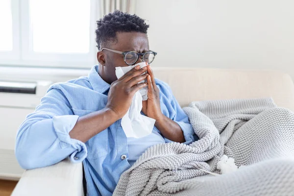 Ill african young woman covered with blanket blowing running nose got fever caught cold sneezing in tissue sit on sofa, sick allergic black girl having allergy symptoms coughing at home, flu conceptIll african young man covered with blanket blowing r