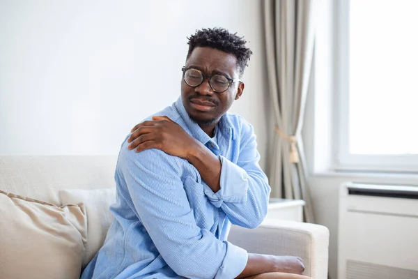 Pain in his shoulder. Upper arm pain, People with body-muscles problem, Healthcare And Medicine concept. Attractive young man sitting on the sofa and holding painful shoulder with another hand.