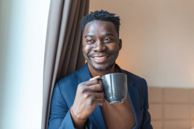İş adamı, otel odasının penceresinde durmuş kahve içiyor. Düşünceli genç Afrikalı iş adamı elinde bir fincan kahveyle pencerenin kenarında durup dışarıya bakıyor..