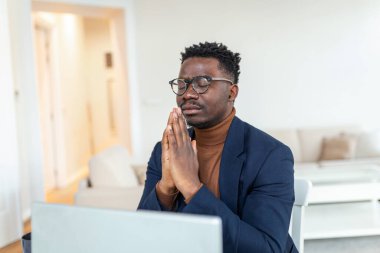 African guy sit at desk with clasped palms closed eyes thinking search problem solution, collect thoughts together, organize his emotional state before important meeting. Prayer hands sign of ask help
