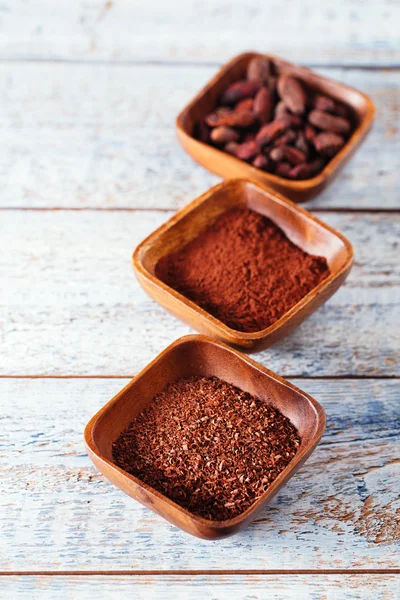 Cocoa beans, powder and grated chocolate in wooden bowls, white — Stock Photo, Image