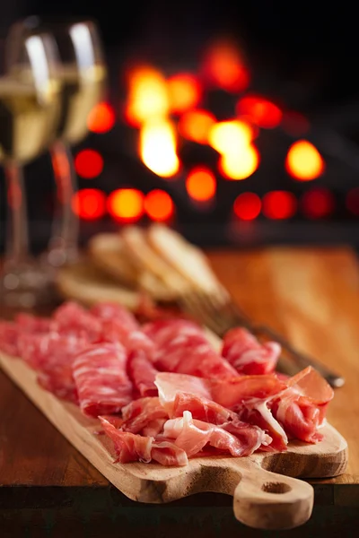 Platter of serrano jamon Cured Meat with cozy fireplace and wine — Stock Photo, Image