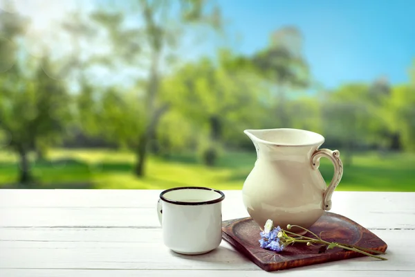 Milchkanne und Becher auf sonnigem Sommerland-Hintergrund — Stockfoto