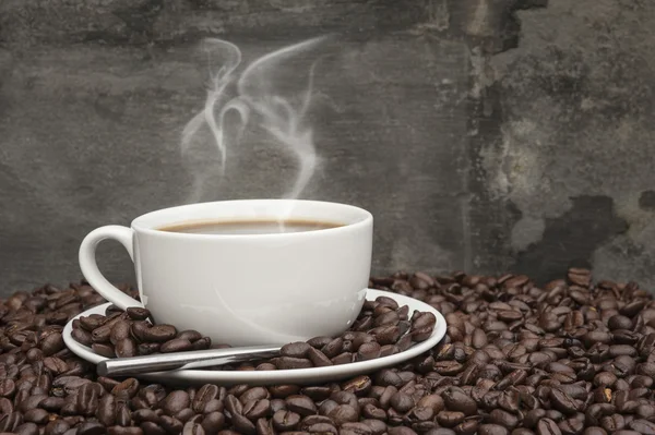 Steaming hot cup of coffee surrounded by dark coffee beans with — Stock Photo, Image