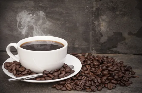 Steaming hot cup of coffee surrounded by dark coffee beans with — Stock Photo, Image