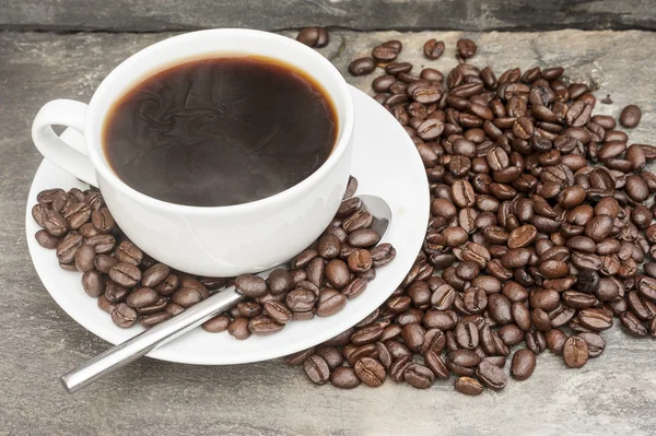 Steaming hot cup of coffee surrounded by dark coffee beans with — Stock Photo, Image