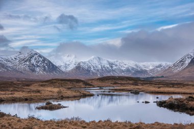 Majestic Winter panorama landscape image of mountain range and peaks viewed from Loch Ba in Scottish Highlands with dramatic clouds overhead