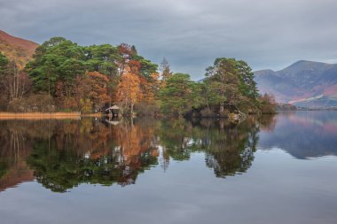 Derwentwater 'daki kayıkhanenin canlı sonbahar gündoğumu sırasında gölün etrafını çevreleyen ormanlık alandaki destansı manzarası.