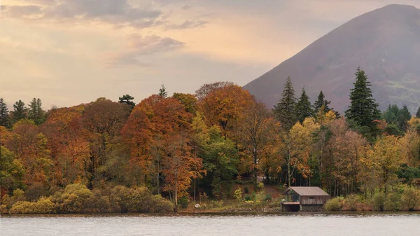 Epic landscape image of boathouse on Derwentwater in Lake District surrounded by vibrant Autumn color woodland