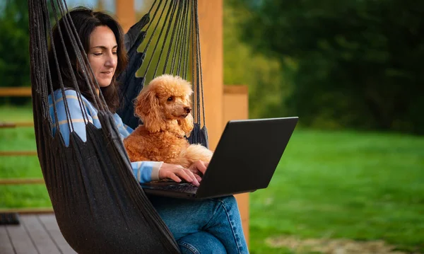 年轻的自由职业者女人带着狗在阳台上挂着的椅子上电脑上工作 在家远程工作的工作场所概念 — 图库照片