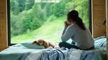 Köpeğiyle yatakta oturan bir kadın pencereden dışarı bakar ve bir dağ görür. Çay ya da kahve içen bir kadın..
