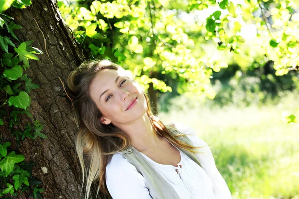Uśmiechający się blond kobieta, opierając się na drzewo — Zdjęcie stockowe