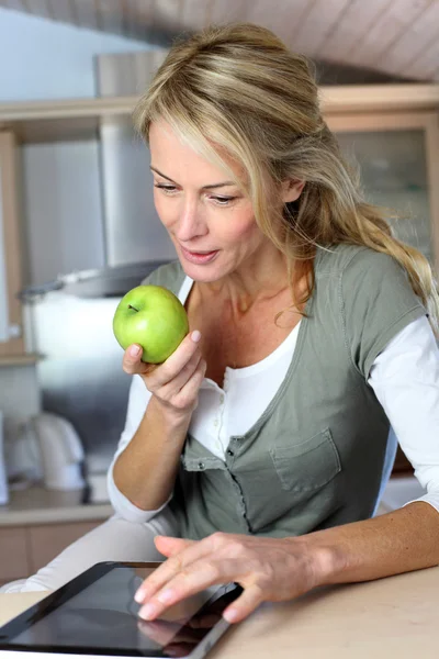 Mulher adulta alegre websurf com tablet e comer maçã — Fotografia de Stock