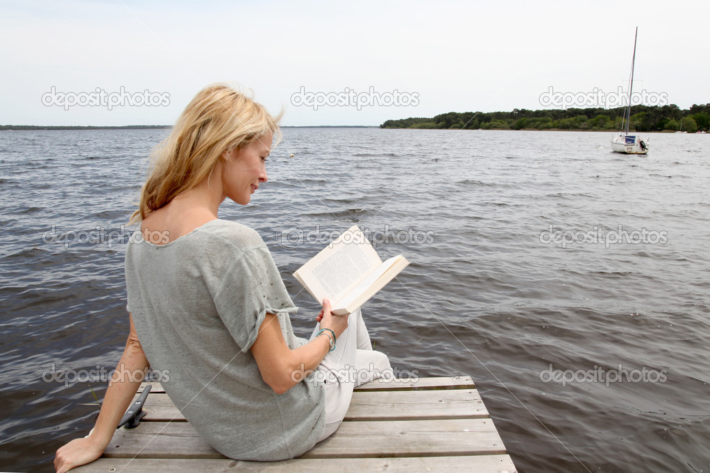 Woman reading book sitting on lake boardwalk