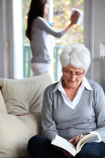 Elderly woman reading book while housekeeper cleans windows — Stock Photo, Image