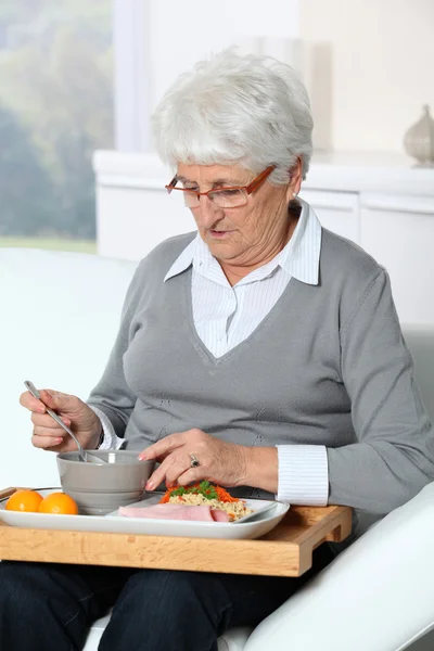 昼食のトレイが付いているソファーに座っている高齢者の女性 — ストック写真