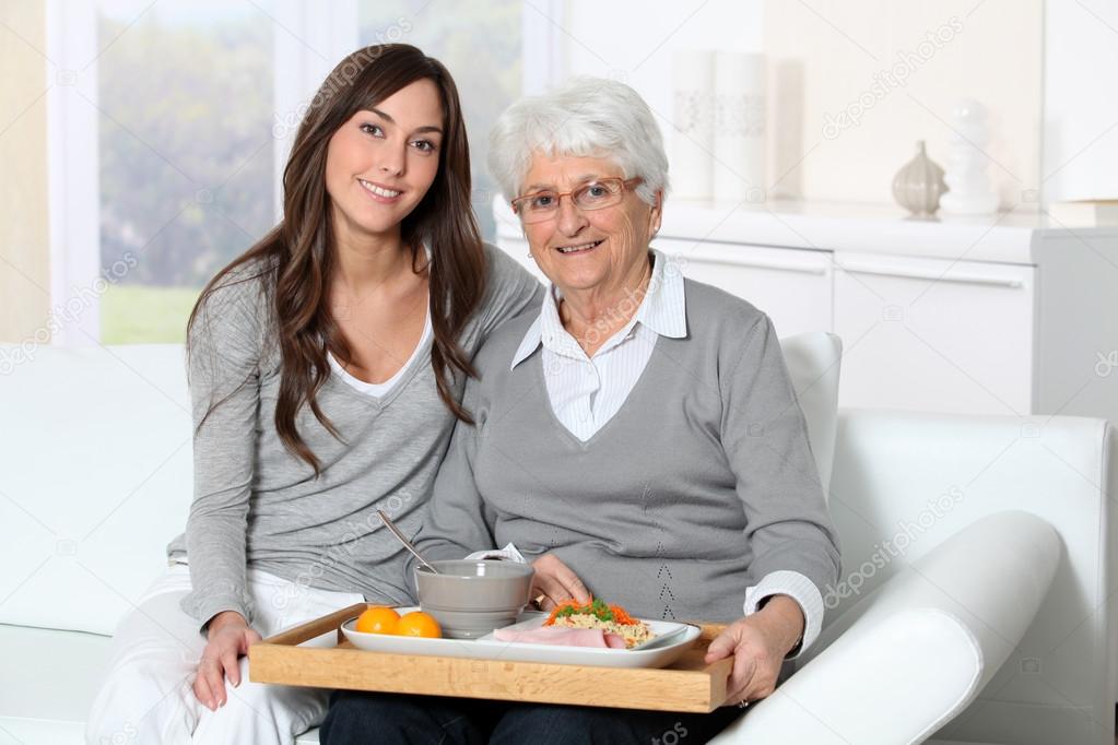 Elderly woman and home carer sitting in sofa with lunch tray
