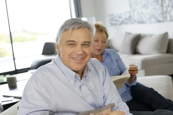Uśmiechający się starszy człowiek siedzi w kanapie, żona w tle — Zdjęcie stockowe