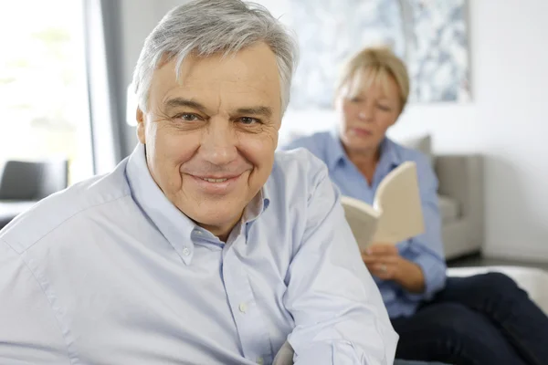 Smiling senior man sitting in couch, wife in background — Stock Photo, Image