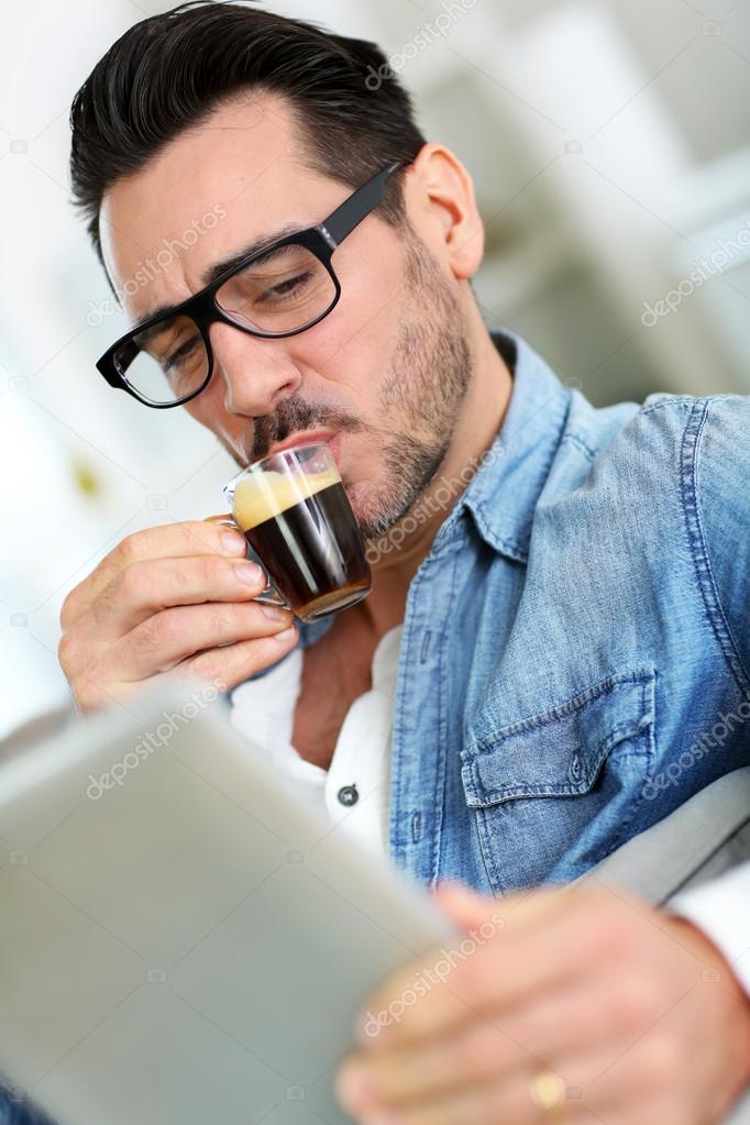Man with glasses drinking coffee and using tablet