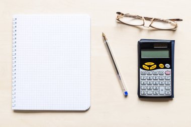 Açık kahverengi ahşap tahtada, boş sayfanın karesi, sarmal defter, kalem, gözlük ve finansal hesap makinesinin üst görünümü