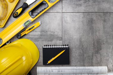 Müteahhit teması. Müteahhidin alet çantası, sarı başlık, libella, el testeresi. Gri fayansların üzerinde planlar ve defter.