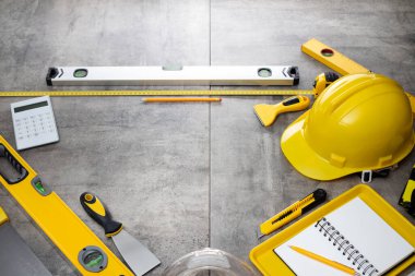 Müteahhit teması. Müteahhidin alet çantası, sarı başlık, libella, el testeresi. Gri fayansların üzerinde planlar ve defter.