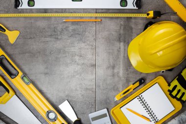 Müteahhit teması. Müteahhidin alet çantası, sarı başlık, libella, el testeresi. Gri fayansların üzerinde planlar ve defter.