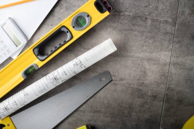 Müteahhit teması. Müteahhidin alet çantası, sarı başlık, libella, el testeresi. Gri fayansların üzerinde planlar ve defter.