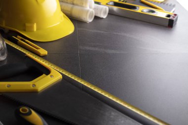 İş kasılma teması. Müteahhidin alet çantası, sarı başlık, libella, el testeresi. Gri fayansların üzerinde planlar ve defter.