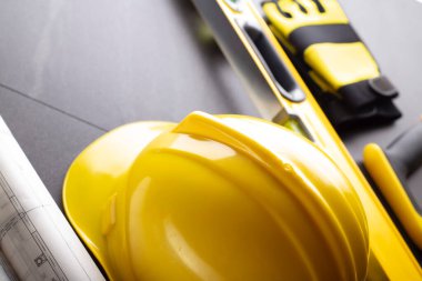 İş kasılma teması. Müteahhidin alet çantası, sarı başlık, libella, el testeresi. Gri fayansların üzerinde planlar ve defter.