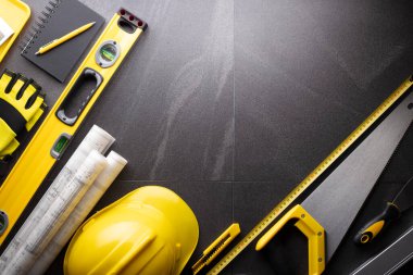 İş kasılma teması. Müteahhidin alet çantası, sarı başlık, libella, el testeresi. Gri fayansların üzerinde planlar ve defter.