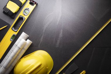 İş kasılma teması. Müteahhidin alet çantası, sarı başlık, libella, el testeresi. Gri fayansların üzerinde planlar ve defter.