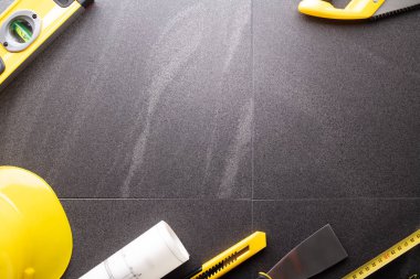 İş kasılma teması. Müteahhidin alet çantası, sarı başlık, libella, el testeresi. Gri fayansların üzerinde planlar ve defter.