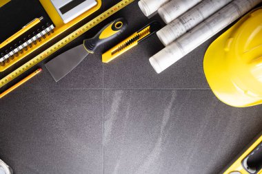 İş kasılma teması. Müteahhidin alet çantası, sarı başlık, libella, el testeresi. Gri fayansların üzerinde planlar ve defter.