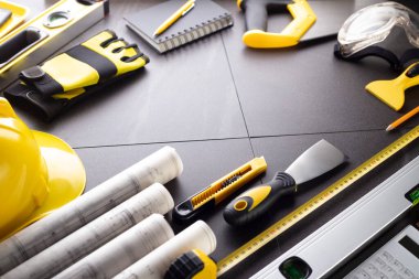 İş kasılma teması. Müteahhidin alet çantası, sarı başlık, libella, el testeresi. Gri fayansların üzerinde planlar ve defter.