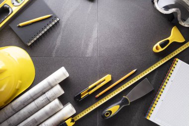 İş kasılma teması. Müteahhidin alet çantası, sarı başlık, libella, el testeresi. Gri fayansların üzerinde planlar ve defter.