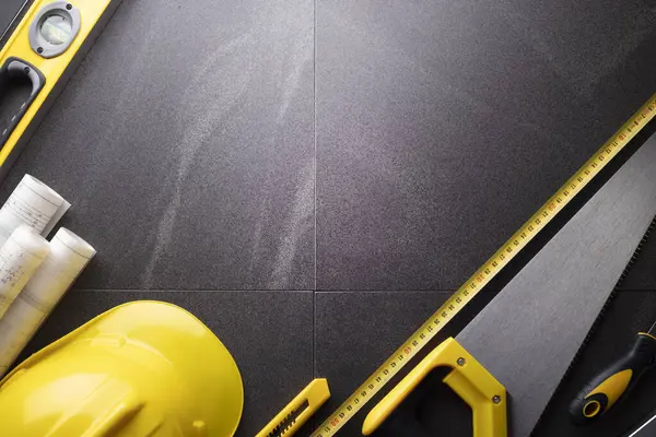 İş kasılma teması. Müteahhidin alet çantası, sarı başlık, libella, el testeresi. Gri fayansların üzerinde planlar ve defter.