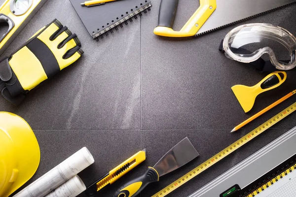 İş kasılma teması. Müteahhidin alet çantası, sarı başlık, libella, el testeresi. Gri fayansların üzerinde planlar ve defter.