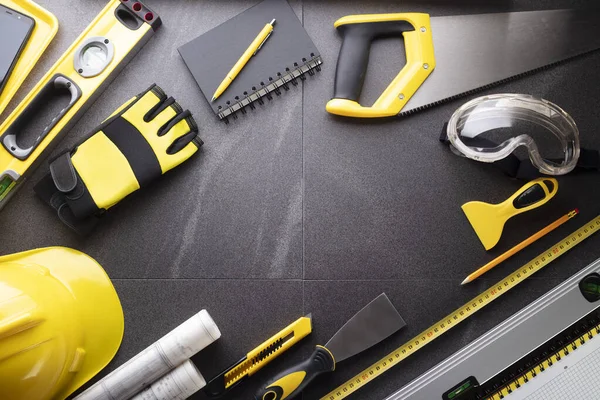 İş kasılma teması. Müteahhidin alet çantası, sarı başlık, libella, el testeresi. Gri fayansların üzerinde planlar ve defter.