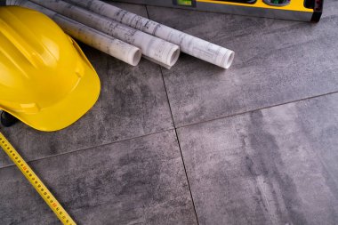 Müteahhit teması. Müteahhidin alet çantası, sarı başlık, libella, el testeresi. Gri fayansların üzerinde planlar ve defter.