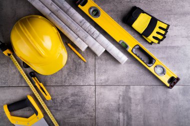 Müteahhit teması. Müteahhidin alet çantası, sarı başlık, libella, el testeresi. Gri fayansların üzerinde planlar ve defter.