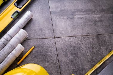 Müteahhit teması. Müteahhidin alet çantası, sarı başlık, libella, el testeresi. Gri fayansların üzerinde planlar ve defter.