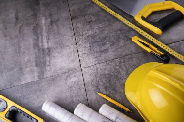 Müteahhit teması. Müteahhidin alet çantası, sarı başlık, libella, el testeresi. Gri fayansların üzerinde planlar ve defter.