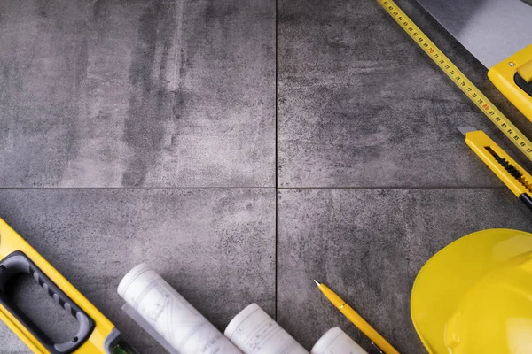 Müteahhit teması. Müteahhidin alet çantası, sarı başlık, libella, el testeresi. Gri fayansların üzerinde planlar ve defter.