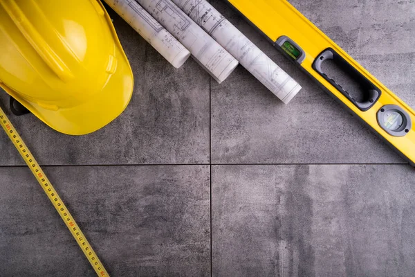 Müteahhit teması. Müteahhidin alet çantası, sarı başlık, libella, el testeresi. Gri fayansların üzerinde planlar ve defter.