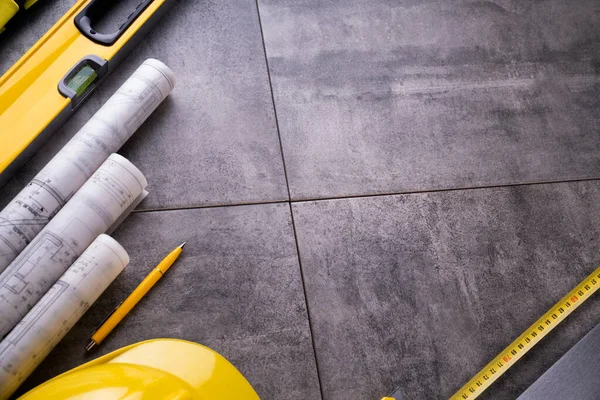 Müteahhit teması. Müteahhidin alet çantası, sarı başlık, libella, el testeresi. Gri fayansların üzerinde planlar ve defter.