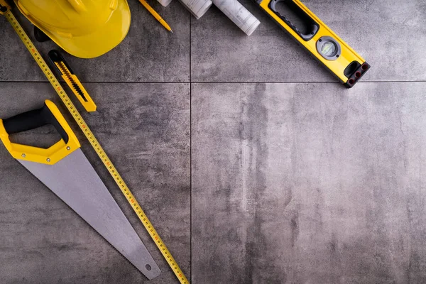 Müteahhit teması. Müteahhidin alet çantası, sarı başlık, libella, el testeresi. Gri fayansların üzerinde planlar ve defter.