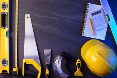 Müteahhit teması. Müteahhitin planları, defter ve alet çantası: sarı baret, iftiracı, gri fayansların arka planında testere ve yarda..